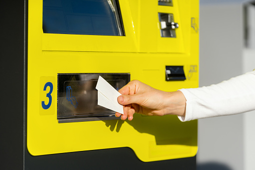 Cropped shot of woman hand holding tickets for public transport against automatic terminal machine. Choosing and buying paper document for travel. Copy space.