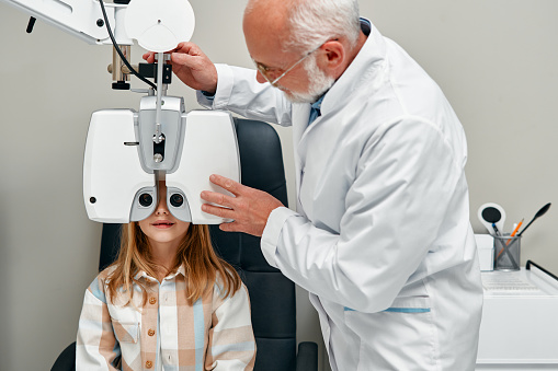 Cute girl child doing eye test checking examination with optometrist in optical shop. Optometrist doing sight testing for child patient in clinic. Ophthalmology and eye correction.