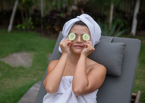 Close-up shot of Asian woman putting cucumber above her eyes to help soothe puffiness and reduce dark circles under the eyes. She's sitting on tanning bed, outside her villa , wearing a towel on her body and head.