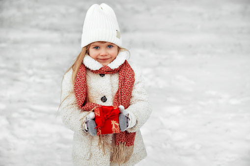 Merry Christmas and Happy New Year. Cute little girl holding a red box with a gift outside in snowy weather. Preparing for the holidays.