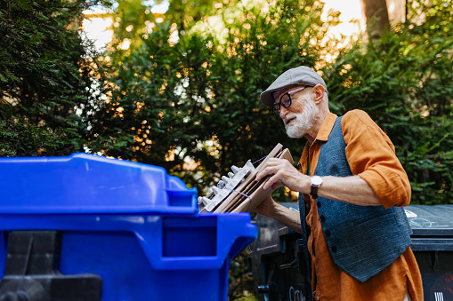 Senior man throwing paper waste and cardboards into recycling container in front apartment. Elderly man sorting the waste according to material into colored bins.