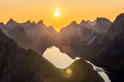 Sunset bathes the jagged peaks of Reinebringen in Lofoten Islands, Norway with golden light, the midnight sun casting a serene glow over the calm waters below, highlighting the contrasts of Arctic landscape