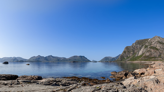Lofoten's rugged coastline unfolds under a clear sky, with mountains rising behind the serene Norwegian Sea, a pristine example of Norway's natural splendor