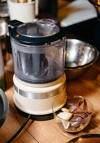 An electric food processor with a clear bowl alongside fresh ingredients including anchovies and garlic cloves.