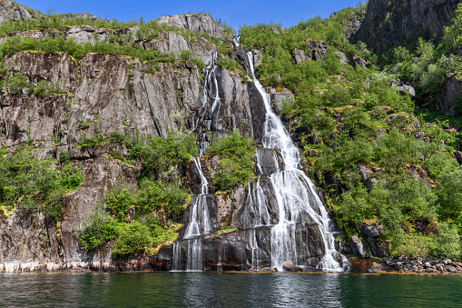 Sunlight dances on the cascading waters of a multi-tiered waterfall in the lush, green setting of Trollfjorden, Lofoten, Norway, a pristine natural spectacle