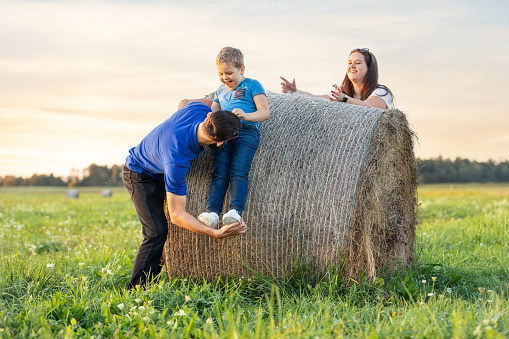 Parents help their little son to climb on a hay bale in a beautiful rural summer meadow.