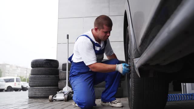 Auto mechanic in blue uniform mounts tire using pneumatic wrench. Professional tire fitting, garage workflow. Skilled worker replaces wheel, car maintenance, repair services. Seasonal tire change.