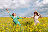 Two young women in beautiful dresses walk in a field with yellow blooming rapeseed.