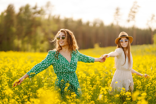 Two young women in beautiful dresses walk in a field with yellow blooming rapeseed. Beautiful girlfriends enjoying the weather, having fun in a flowering field. Concept of fun, relaxation.