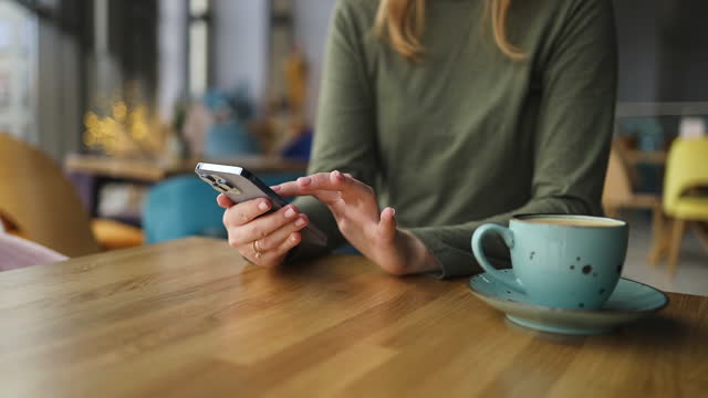 Female hands holding smartphone, drinking and enjoying coffee on wooden table at coffee shop cafe. close up.