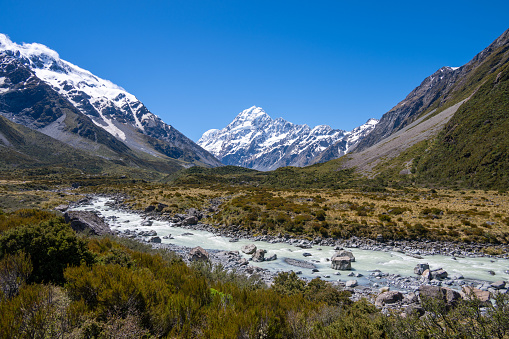 Discover the picturesque landscape in New Zealand's South Island leading to the renowned Mount Cook National Park. Behold the stunning sight of snow-capped Mount Cook, majestically framed by other towering mountain peaks. In front a glacier river.