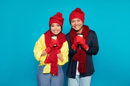 Smiling young asian family couple in hats and scarves warming up with cups of hot drink takeaway, isolated on blue studio background. Couple enjoy weekend, holidays, winter and New Year, Xmas