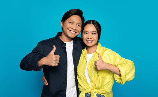 Positive young japanese guy and woman in casual show thumb up sign with hand, isolated on blue studio background. Emotions recommendation, approve gesture, good news sign