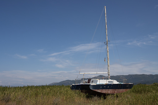 Old rusted abandoned yacht on the filed with a high grass. Mountains in the background. Blue sky with light white clouds in the spring. Roda, Corfu, Greece.