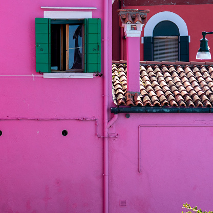 A vibrant pink building with green shutters and an open window sits atop a traditional brown roof