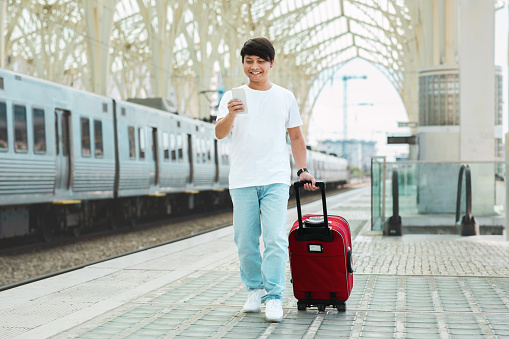 Asian guy in casual outfit tourist walking by train station, carrying red luggage, using smartphone, checking train schedule, buying ticket online, make taxi reservation on mobile app, copy space