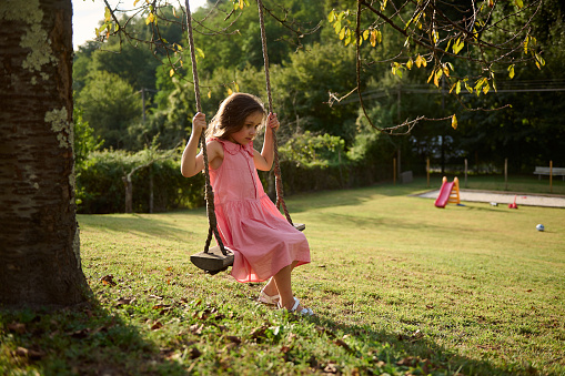 Little Caucasian child girl in pink dress, swinging on a wooden rustic swing outdoor at sunset. Kids entertainment. People. Hobbies and leisures