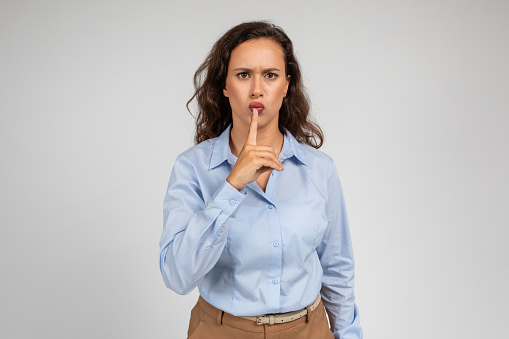 Strict sad angry young european businesswoman put finger to lips, make shhh sign with hand, isolated on gray studio background. Secret news, gossip and emotions, ad and offer