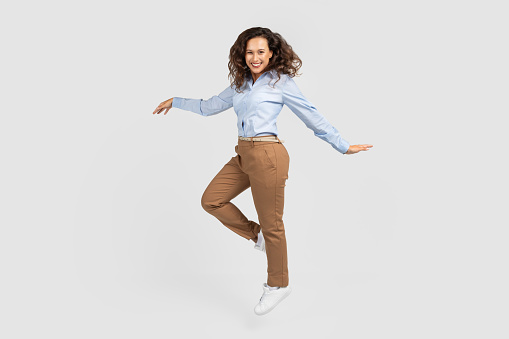 Happy young european lady enjoy win, good mood, jump, freeze in air, joy and triumph, celebrate success and dance in work, business, isolated on gray studio background, full length