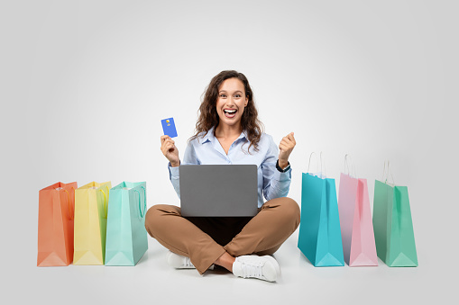 European happy shocked woman with laptop and credit card sit on floor with many bags with purchases, isolated on gray studio background. Work, business remotely, sale for shopaholic
