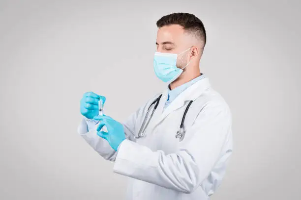 Concentrated male doctor in a surgical mask and gloves carefully preparing a syringe for vaccination, epitomizing healthcare and patient safety