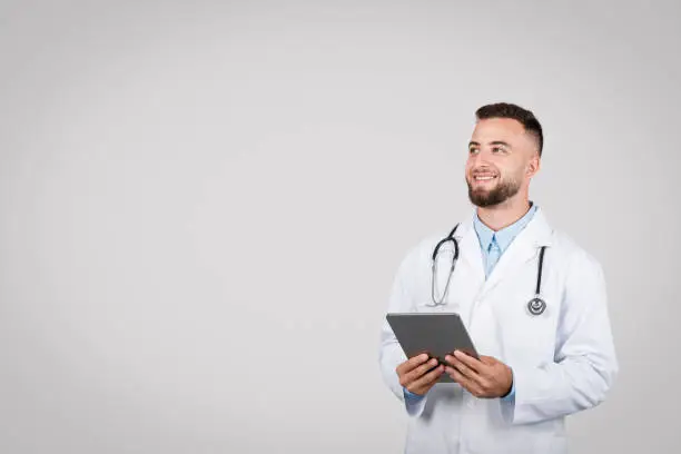 European young male doctor in white coat holding digital tablet, gazing at copy space over simple grey background, exemplifying modern healthcare