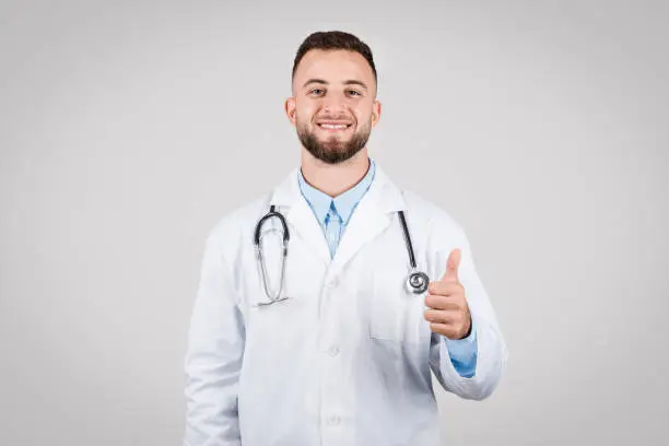 Cheerful European young male doctor in white coat, giving thumb up and smiling, expressing positivity and confidence in healthcare, set against grey background