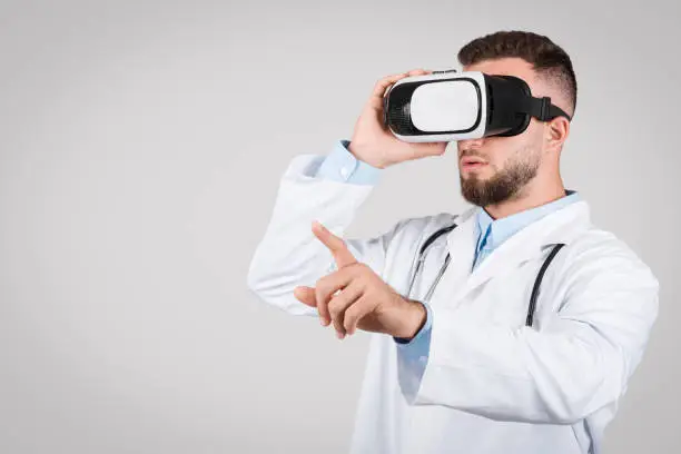 European young male doctor dressed in white coat, wearing virtual reality glasses, representing futuristic medical technology, against plain grey background