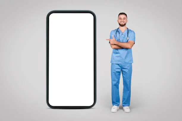 European young male doctor in blue coat standing beside huge cellphone screen, pointing at its blank screen, ideal for medical info display, against grey backdrop