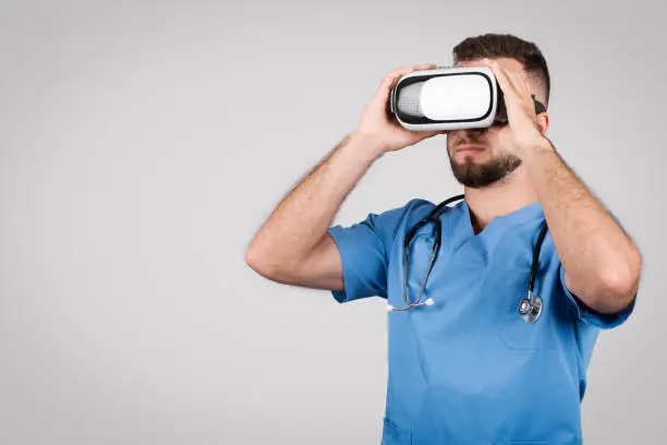 European young male doctor in blue coat, immersed in using virtual reality glasses, representing modern technologies in medicine, against grey backdrop, copy space