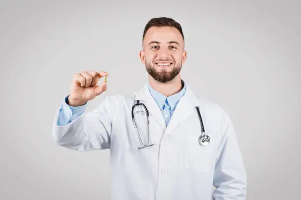 European young male doctor in white coat, cheerfully presenting pill, symbolizing positive healthcare solutions, set against simple grey background for medical ads