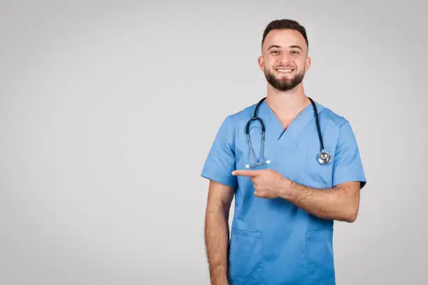 Young European male doctor in blue coat, pointing to empty space at the side, perfect for medical advertising or information, set against grey background