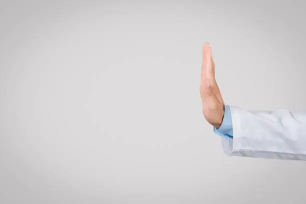 European young male doctor in white coat, displaying stop hand gesture, side view, signifying caution or warning, against simple grey background, free space