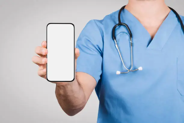 European young male doctor in blue coat, displaying cellphone with white blank screen, ideal for medical advertising content, set against grey background, mockup