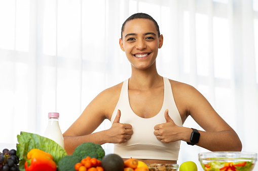 Smiling millennial latin woman in sportswear make thumb up sign with hands in kitchen interior, recommends fresh vegetables, salad. Diet fit, weight loss, healthy food and lifestyle approve at home