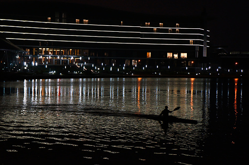 A Man Rowing a Kayak on the River in a City at Night by Modern Buidings and Color Reflections on Water.