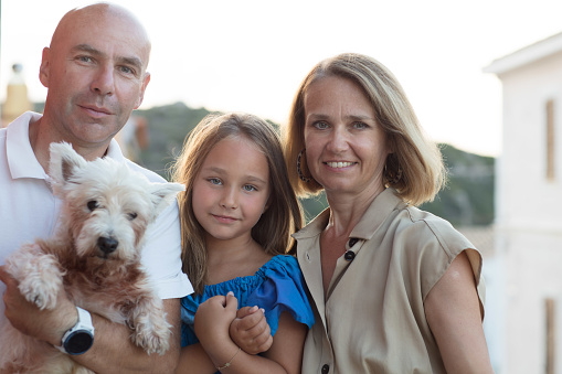 Close-up portrait of a family, consisting of mum, dad, a little girl and a small dog. They stand at dusk on a balcony in the town of Santa Teresa Gallura, Sardinia.