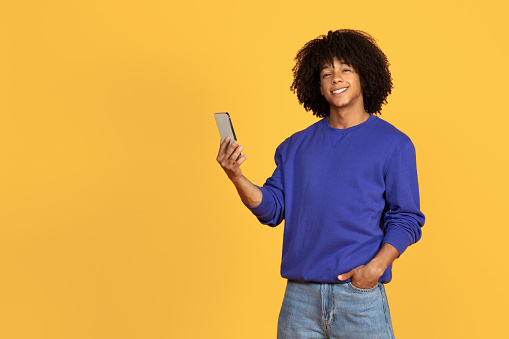Happy Young Black Guy Using Mobile Phone And Smiling At Camera, Cheerful Millennial African American Man With Smartphone In Hand Standing Isolated Over Yellow Studio Background, Copy Space