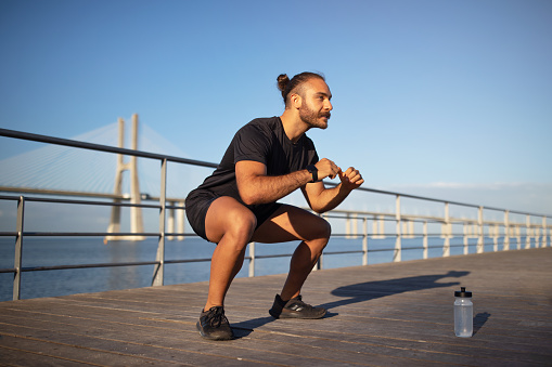 Positive young caucasian man enjoys outdoor fitness workout while squats at sea pier. Athletic guy having sports on weekend morning. Active lifestyle and body care, weight loss