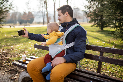 Beard handsome man with his baby boy son in a baby carrier is sitting on the park bench and making a selfie. Love emotion and strong connection.