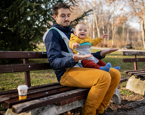 Young handsome man with his baby boy son in a baby carrier is sitting on the park bench and drinking coffee. They are playing and laughing together. Love emotion. Strong connection between father and son.