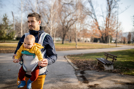 Young handsome man with his baby boy son in a baby carrier is walking through the public park. They are enjoying the cold but sunny day. Positive emotion and healthy life.