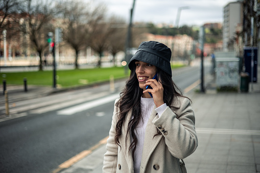 copy space Caucasian brunette girl with hat and coat talking on cell phone walking through the city after work