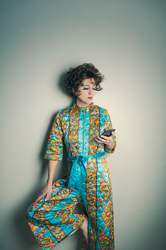 A strange image of a vintage fashion model dressed in a late 1960s paisley romper posing as she looks at a modern day cell phone.