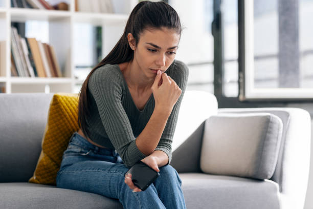 istock Depressed young woman thinking about her problems while holding mobile phone sitting on the sofa at home. 1915553306