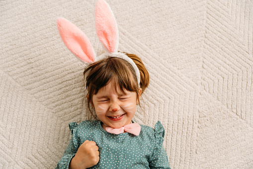 Cheerful little girl  with face painting as a Easer bunny, lying on the carpet, smiling and looking at camera