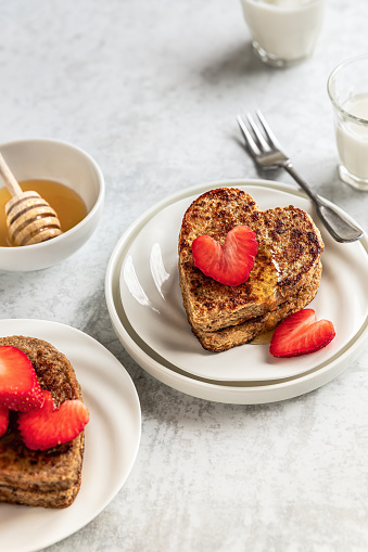 Whole grain caramelized bread slices in heart shape topped with fresh strawberries and melted honey; French toasts on plates ready to eat; romantic breakfast for two;