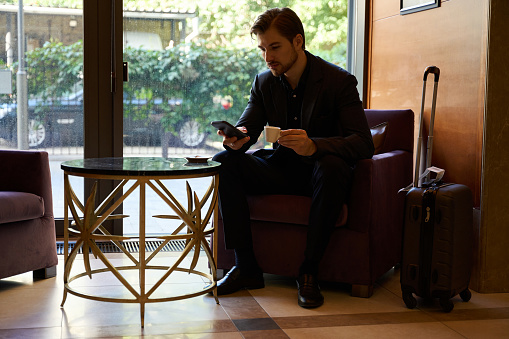 Young man sitting on armchair at lobby bar while watching something on smartphone during coffeetime
