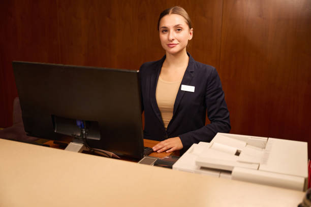 Woman receptionist standing in front of computer at reception counter in hotel while waiting for visitors