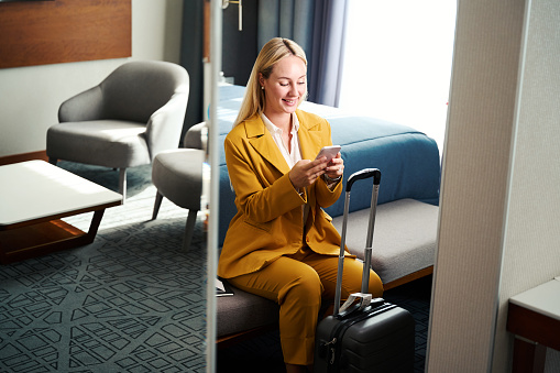 Smiling young businesswoman texting on phone while sitting on ottoman bench in hotel room